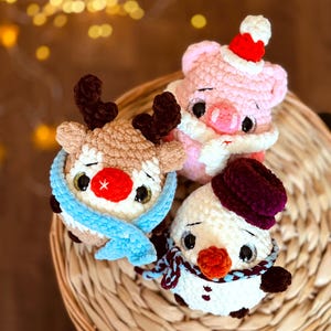 Könnte beinhalten: Drei gehäkelte Weihnachtsornamente: ein Rentier mit einem blauen Schal, ein Schneemann mit einem burgunderfarbenen Hut und Schal und ein rosa Schwein mit einem Weihnachtsmann-Hut.