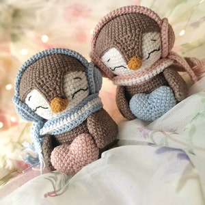 Op de afbeelding: Twee bruine gehaakte pingu&iuml;n speelgoedjes die blauwe en roze sjaals en oorwarmers dragen. Elke pingu&iuml;n houdt een hartvormig gehaakt voorwerp vast, een roze en een blauwe.