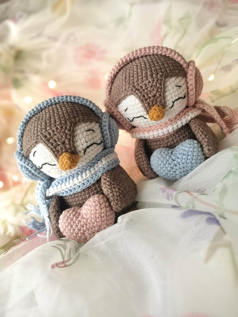 Op de afbeelding: Twee bruine gehaakte pingu&iuml;n speelgoedjes die blauwe en roze sjaals en oorwarmers dragen. Elke pingu&iuml;n houdt een hartvormig gehaakt voorwerp vast, een roze en een blauwe.