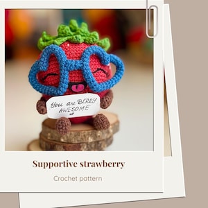May include: A crocheted strawberry character with blue heart-shaped glasses and a green leafy top. The strawberry holds a sign that reads "You are BERRY AWESOME." The image is set on a wooden base, with the words "Supportive strawberry" and "Crochet pattern" below.
