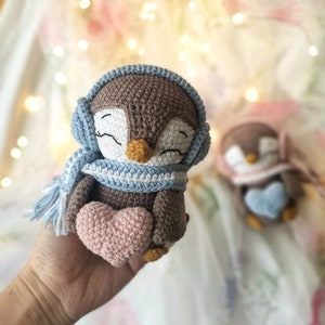 Op de afbeelding: Een bruine gehaakte pingu&iuml;n met een blauw-witte sjaal en een blauwe gehaakte muts. De pingu&iuml;n houdt een roze gehaakt hart vast.