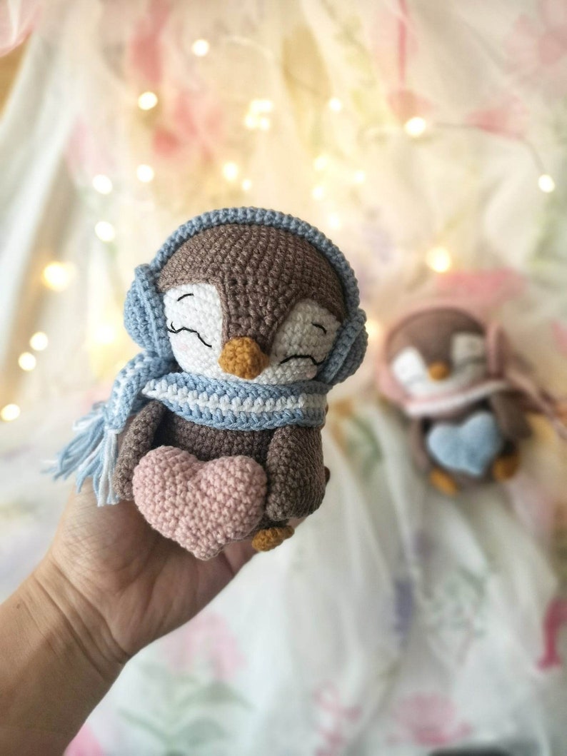 Op de afbeelding: Een bruine gehaakte pingu&iuml;n met een blauw-witte sjaal en een blauwe gehaakte muts. De pingu&iuml;n houdt een roze gehaakt hart vast.