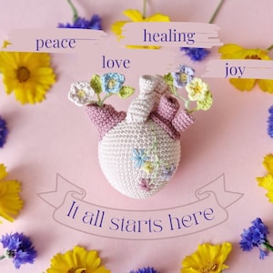 Peut inclure: Décoration en forme de cœur crochetée avec des accents floraux et le texte « It all starts here ». Le cœur est entouré de fleurs jaunes et bleues, avec les mots « paix », « guérison », « amour » et « joie » au-dessus.