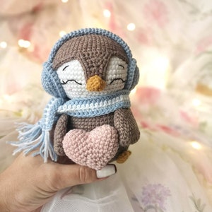 Op de afbeelding: Een gehaakt pingu&iuml;n speelgoed met een blauwe sjaal en oorkappen, die een roze gehaakt hart vasthoudt.