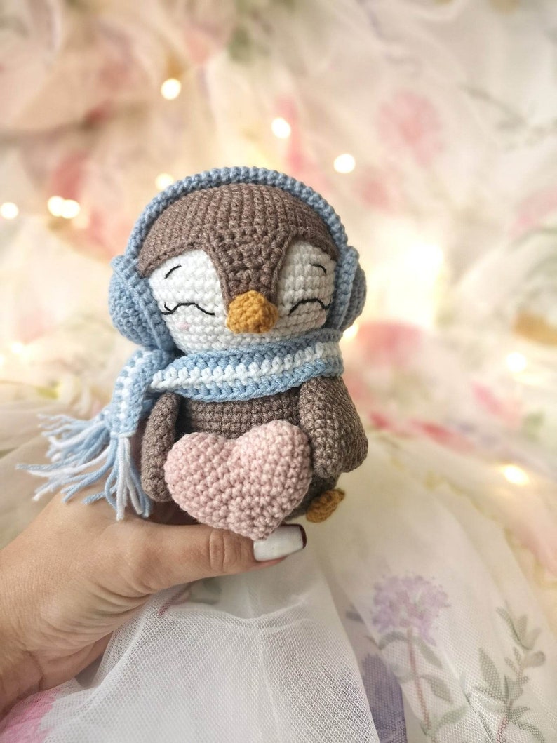 Op de afbeelding: Een gehaakt pingu&iuml;n speelgoed met een blauwe sjaal en oorkappen, die een roze gehaakt hart vasthoudt.