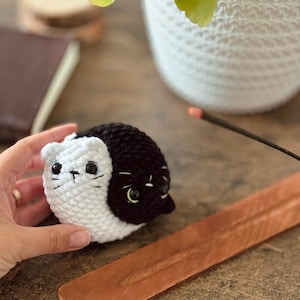 Pode incluir: Um brinquedo de gato de croch&ecirc; preto e branco com olhos pretos e nariz branco. O gato est&aacute; sentado em uma superf&iacute;cie de madeira.