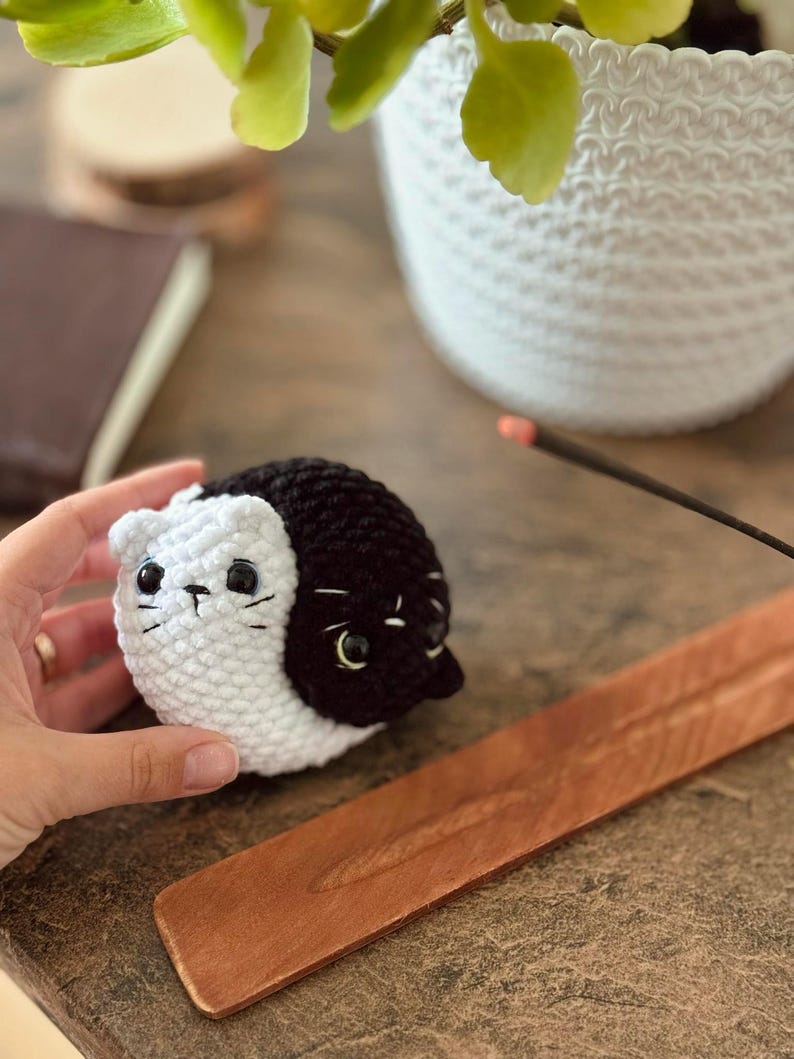 Pode incluir: Um brinquedo de gato de croch&ecirc; preto e branco com olhos pretos e nariz branco. O gato est&aacute; sentado em uma superf&iacute;cie de madeira.