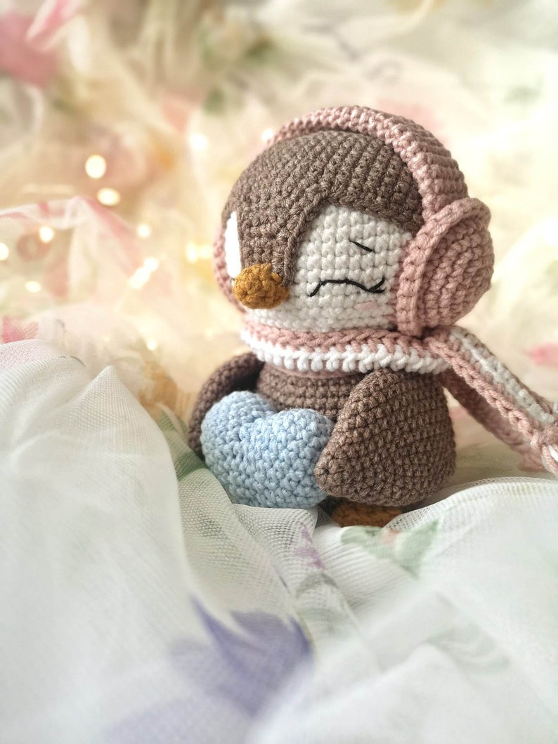 Op de afbeelding: Een bruine gehaakte pingu&iuml;n met roze koptelefoon en een sjaal, die een lichtblauw gehaakt hart vasthoudt. De pingu&iuml;n zit op een witte en roze stoffen achtergrond.