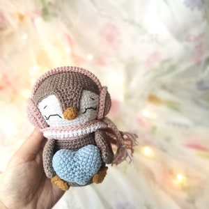 Op de afbeelding: Een bruine gehaakte pingu&iuml;n met een roze en witte sjaal en een roze muts met oorflappen. De pingu&iuml;n houdt een lichtblauw gehaakt hart vast.