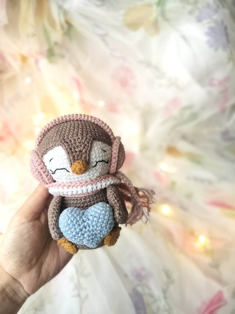 Op de afbeelding: Een bruine gehaakte pingu&iuml;n met een roze en witte sjaal en een roze muts met oorflappen. De pingu&iuml;n houdt een lichtblauw gehaakt hart vast.