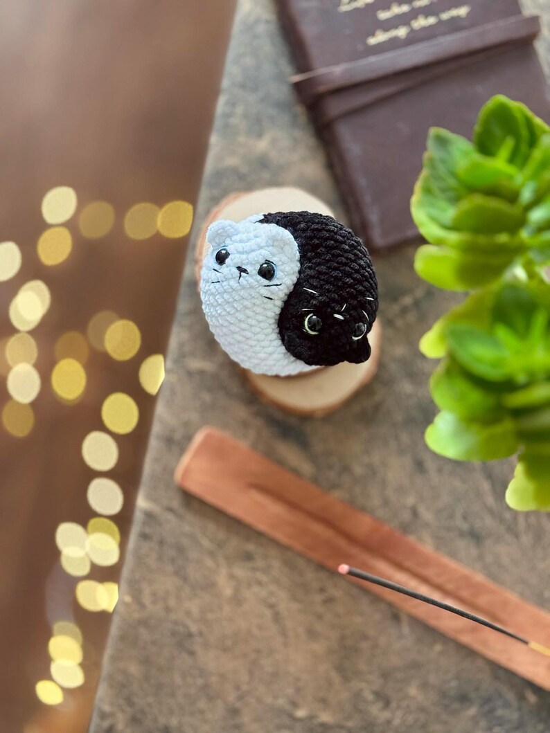 Pode incluir: Uma figura de gato yin yang de croch&ecirc; feita com fio preto e branco. O gato est&aacute; sentado em uma base de madeira.