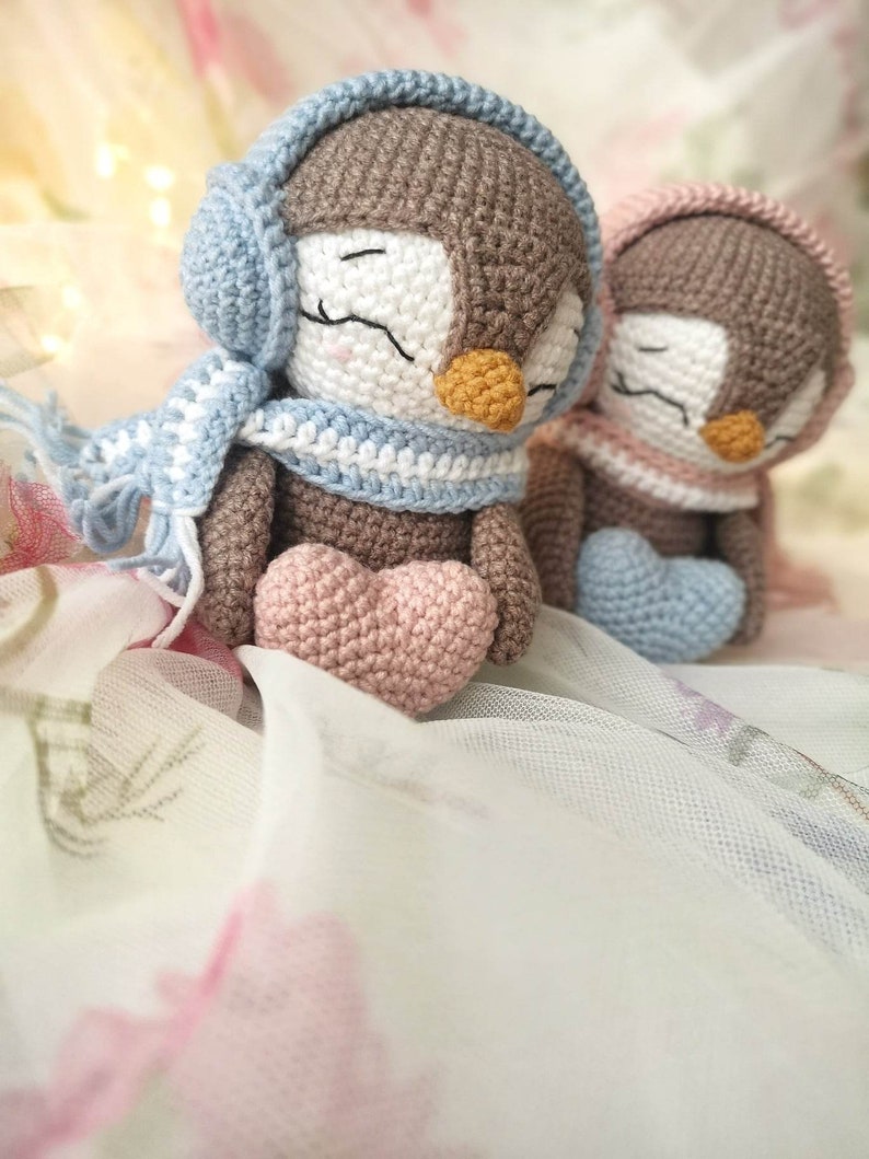 Op de afbeelding: Twee gehaakte pingu&iuml;n speelgoedjes die blauwe en roze oorkappen en sjaals dragen. De pingu&iuml;ns houden roze en blauwe harten vast.