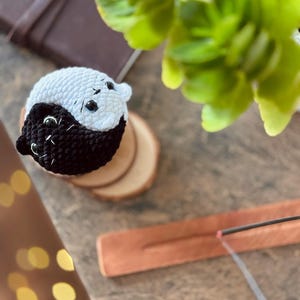 Pode incluir: Uma figura de gato yin yang de croch&ecirc; preto e branco em uma base de madeira. O gato est&aacute; sentado em um porta-incenso de madeira com um bast&atilde;o de incenso aceso.