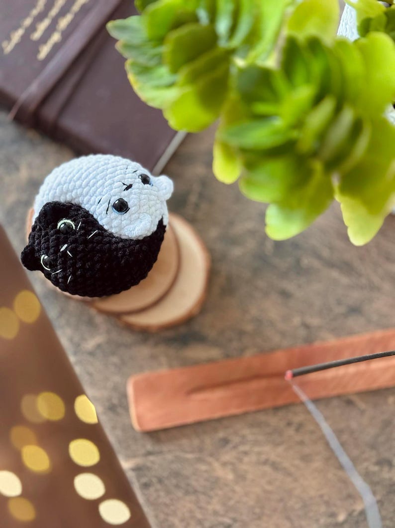 Pode incluir: Uma figura de gato yin yang de croch&ecirc; preto e branco em uma base de madeira. O gato est&aacute; sentado em um porta-incenso de madeira com um bast&atilde;o de incenso aceso.