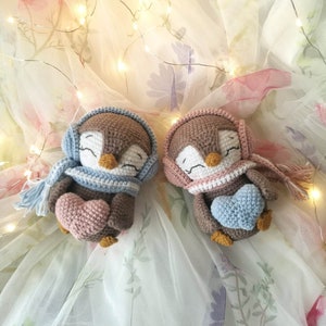Op de afbeelding: Twee gehaakte pingu&iuml;n knuffels die roze en blauwe harten vasthouden. De pingu&iuml;ns dragen sjaals en hebben bruine ogen. De knuffels liggen op een witte tule achtergrond met bloemenpatronen.