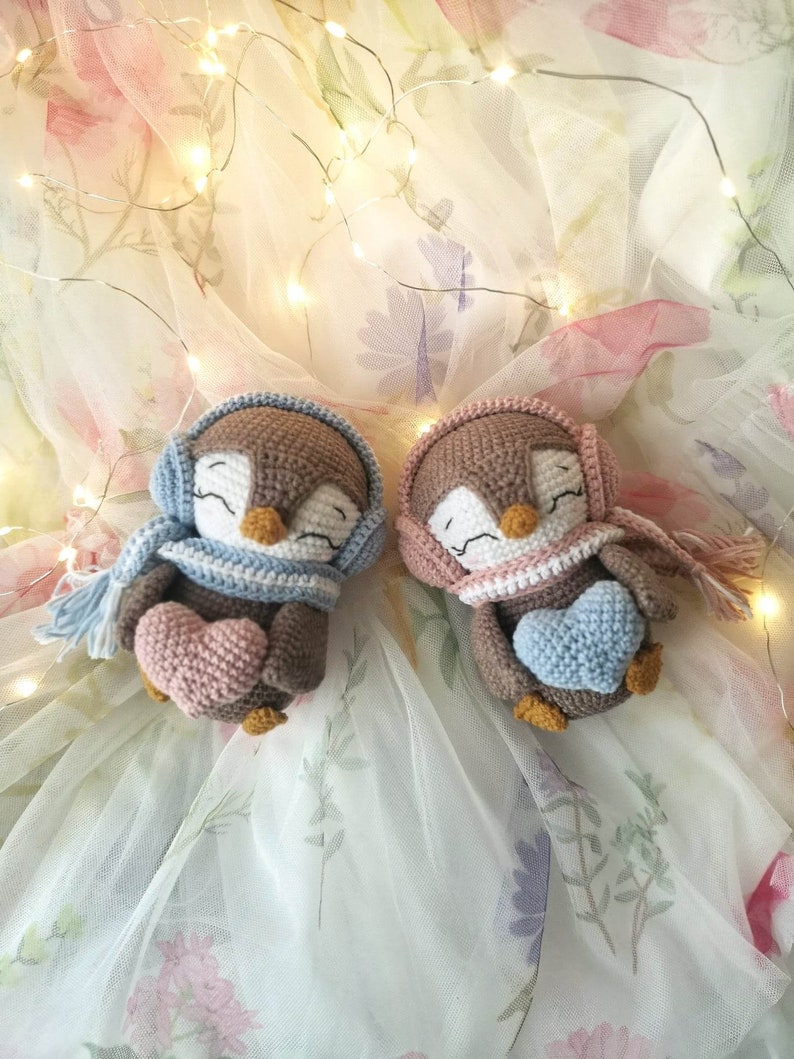 Op de afbeelding: Twee gehaakte pingu&iuml;n knuffels die roze en blauwe harten vasthouden. De pingu&iuml;ns dragen sjaals en hebben bruine ogen. De knuffels liggen op een witte tule achtergrond met bloemenpatronen.