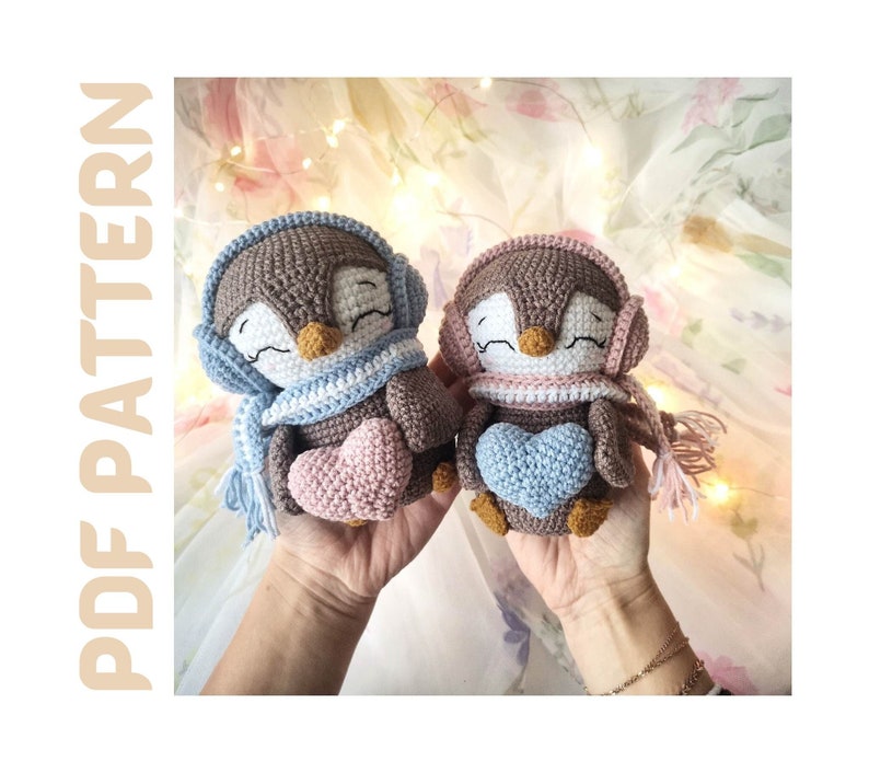 Op de afbeelding: Twee gehaakte amigurumi-pinguinspeelgoedjes met bruine lichamen, witte en blauwe sjaals en roze en blauwe harten. De pingu&iuml;ns houden de harten in hun flippers.