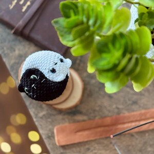 Pode incluir: Um s&iacute;mbolo yin yang de croch&ecirc; feito com fio preto e branco. O s&iacute;mbolo est&aacute; em um descanso de madeira.