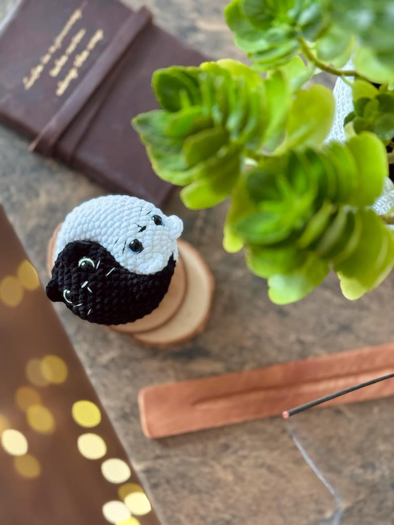 Pode incluir: Um s&iacute;mbolo yin yang de croch&ecirc; feito com fio preto e branco. O s&iacute;mbolo est&aacute; em um descanso de madeira.