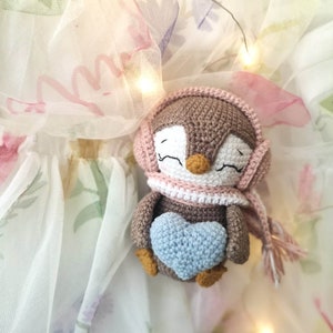 Op de afbeelding: Een bruine gehaakte pingu&iuml;n met een roze gehaakte muts en sjaal, die een blauw gehaakt hart vasthoudt. De pingu&iuml;n zit op een witte tule stof met bloemenpatronen.