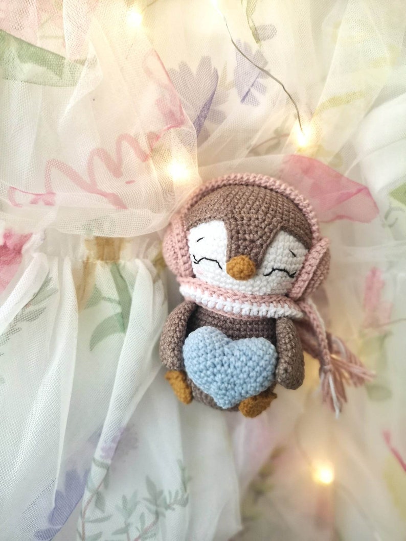 Op de afbeelding: Een bruine gehaakte pingu&iuml;n met een roze gehaakte muts en sjaal, die een blauw gehaakt hart vasthoudt. De pingu&iuml;n zit op een witte tule stof met bloemenpatronen.