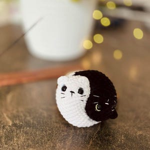 Pode incluir: Um gato de peluche de croch&ecirc; com um design yin yang preto e branco. O gato tem olhos pretos e um nariz branco.
