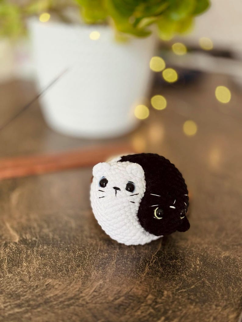 Pode incluir: Um gato de peluche de croch&ecirc; com um design yin yang preto e branco. O gato tem olhos pretos e um nariz branco.