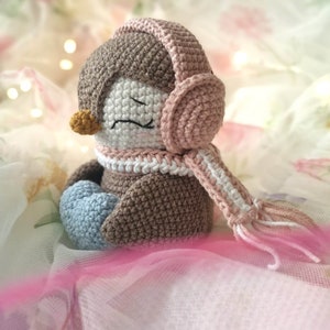 Op de afbeelding: Een gehaakte pingu&iuml;n met roze oordopjes en een witte en roze sjaal. De pingu&iuml;n zit op een witte en roze stoffen achtergrond.