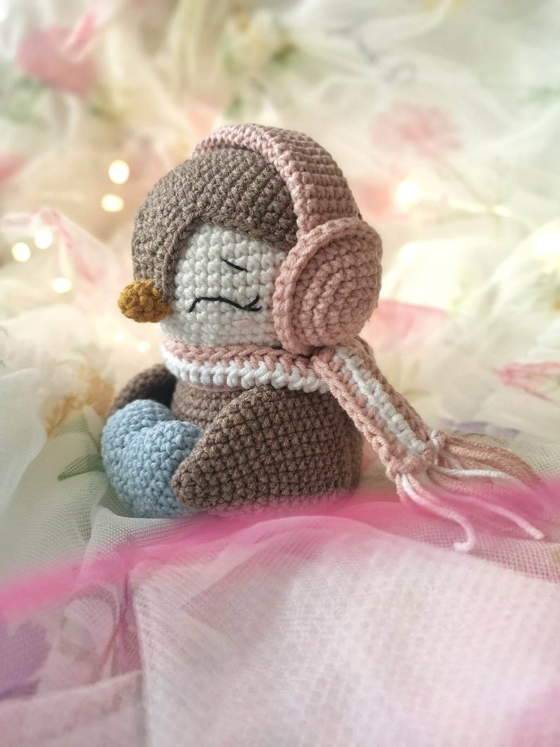 Op de afbeelding: Een gehaakte pingu&iuml;n met roze oordopjes en een witte en roze sjaal. De pingu&iuml;n zit op een witte en roze stoffen achtergrond.
