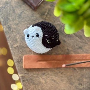 Pode incluir: Um brinquedo de gato de croch&ecirc; com pelo preto e branco. O gato tem olhos pretos e um nariz branco. O brinquedo est&aacute; sentado em uma superf&iacute;cie de madeira.