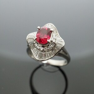 May include: A silver ring with a large red ruby gemstone surrounded by smaller diamonds. The ring has a delicate, intricate design.