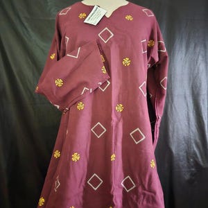 May include: A maroon dress with long sleeves and a V-neck. The dress is adorned with white diamond shapes and yellow floral embroidery. A tag with text hangs near the neckline. The dress is displayed on a mannequin.