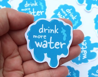 May include: A hand holding a blue sticker with white text that reads "drink more water". The sticker is shaped like a water droplet and has a dripping effect.