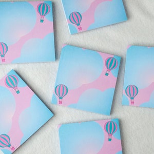 May include: A set of eight pastel blue and pink square sticky notes with a hot air balloon design. Each note features a pink and blue hot air balloon with a teal basket.