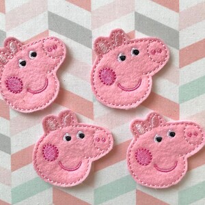 May include: Four pink felt fabric appliques shaped like the head of a cartoon pig. Each piece features embroidered details including two eyes, a snout with two nostrils, a smiling mouth, two ears, and a circular cheek patch in a darker pink thread.