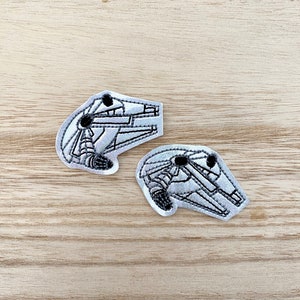Peut inclure: Deux écussons brodés représentant un vaisseau spatial gris et blanc. Les écussons ont un contour détaillé et sont posés sur une surface en bois clair. Ces écussons sont probablement destinés aux vêtements ou aux accessoires.