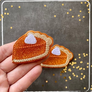 May include: Two embroidered pumpkin pie slice patches featuring a vibrant orange glitter fabric, a light purple dollop of whipped cream, and a tan stitched crust edge. The patches are shaped like triangular pie slices with scalloped crusts.