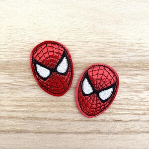 May include: Two oval-shaped embroidered patches featuring the red and black webbed mask design of a superhero character. Each patch displays large, white, almond-shaped eyes outlined in black thread, set against a vibrant red background.