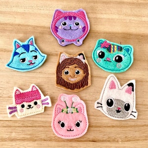 May include: A collection of seven colorful felt cat-shaped appliques. Each cat has a unique design, featuring different colors like blue, purple, pink, and white, with embroidered details. The appliques are arranged on a light brown wooden surface.