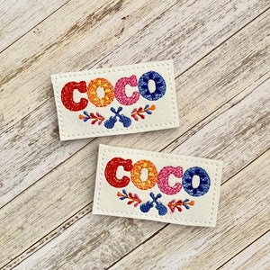 May include: Two rectangular patches with the word "COCO" embroidered in colorful thread. The letters are red, orange, pink, and blue, with floral and musical instrument accents. The patches are on a cream-colored background.