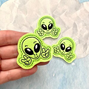 May include: Three lime green alien patches with black eyes and peace signs. The patches are made of felt and have a stitched border. One patch is held in a hand.