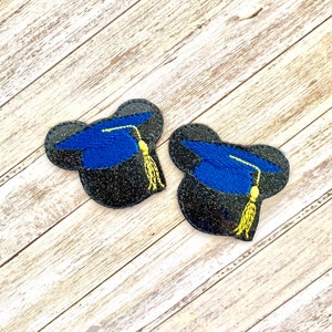 May include: Two embroidered graduation cap patches. Each patch features a blue cap with a yellow tassel, set against a black, glittery background. The patches are shaped like Mickey Mouse heads, adding a playful touch to the graduation theme.