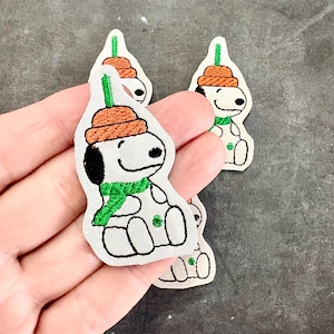 May include: Three embroidered fabric patches featuring a white cartoon dog with a black ear, wearing an orange beanie with a green stem on top and a green scarf. The dog has a small green clover symbol on its chest and is outlined in black thread.