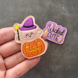 May include: Two embroidered patches. One patch features a light-skinned character wearing a purple witch hat, holding a yellow star wand, and sitting inside an orange pumpkin. The second patch is purple with glitter and the text Wicked CUTE.