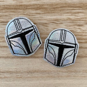 May include: Two silver Mandalorian helmet patches with black outlines and details. The patches have a holographic, glittery finish, and are set against a light wood grain background. The helmets are a popular item from the Star Wars franchise.