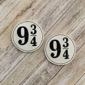 May include: Two round, off-white patches with the number "9 3/4" in black, encircled by a black stitched border. The patches are on a light wood-grain surface. The patches are likely for a Harry Potter theme.