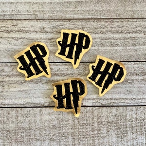 May include: Four black and gold Harry Potter logo cutouts with the letters "HP" and a lightning bolt. The cutouts are on a light wood grain surface.
