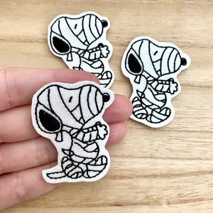 May include: Three embroidered patches featuring a cartoon dog wrapped in white bandages like a mummy. Each patch has a white background with black outlines and details, showing the character with a black ear and nose, standing in a pose with arms held.