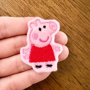 May include: A pink felt applique patch featuring the character Peppa Pig. The character is depicted with a light pink face, a bright red dress, black shoes, and embroidered facial features including eyes, a smile, and a pink cheek detail.