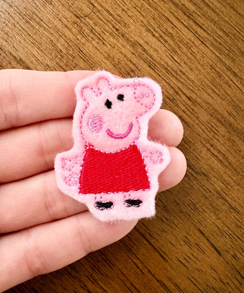 May include: A pink felt applique patch featuring the character Peppa Pig. The character is depicted with a light pink face, a bright red dress, black shoes, and embroidered facial features including eyes, a smile, and a pink cheek detail.